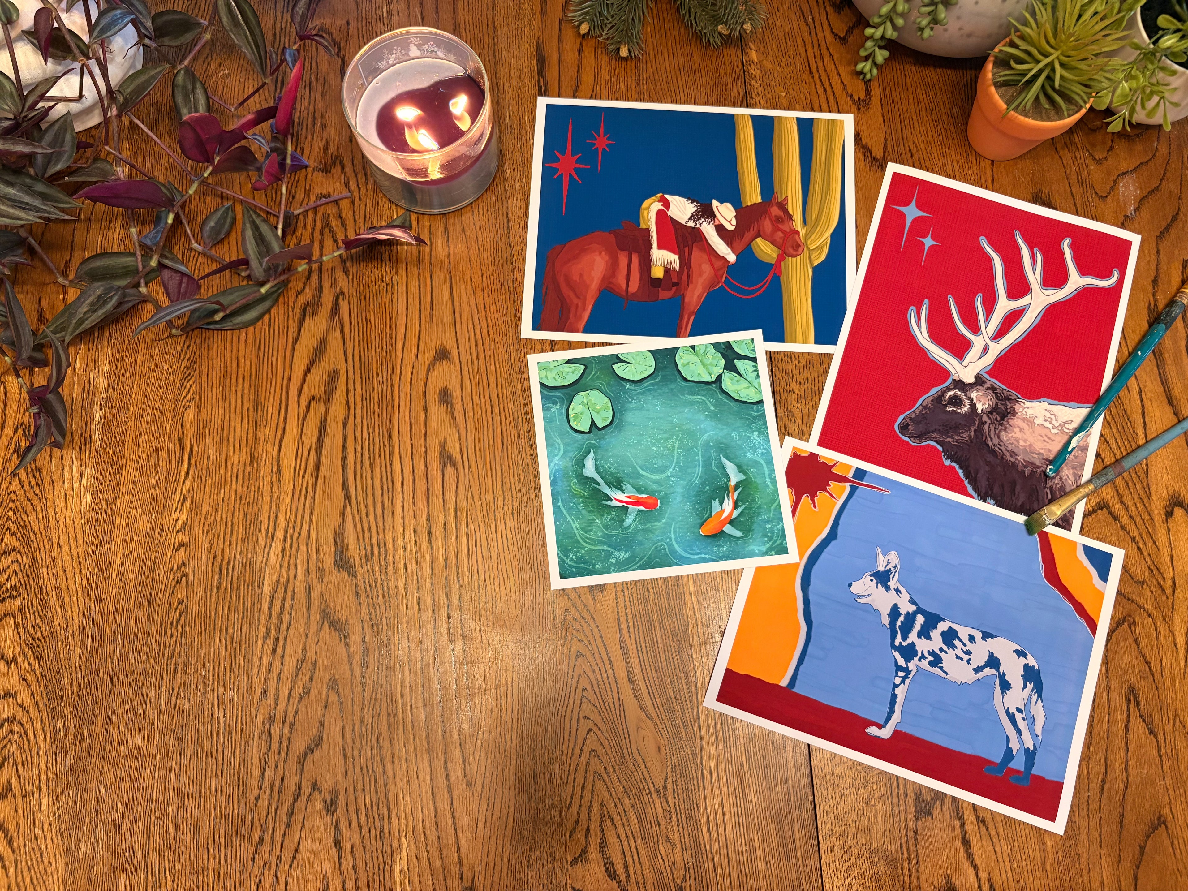 Four colorful art cards featuring a cowboy on a horse, koi fish, an elk, and a spotted dog, arranged on a wooden table next to a lit candle, plants, and paintbrushes.
